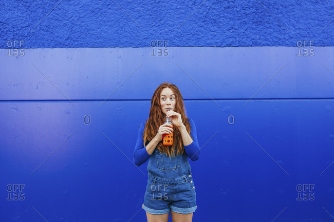 Young woman in blue casual clothing drinking juice while standing against blue wall