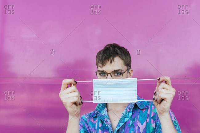Non-binary person holding protective face mask while standing against pink wall