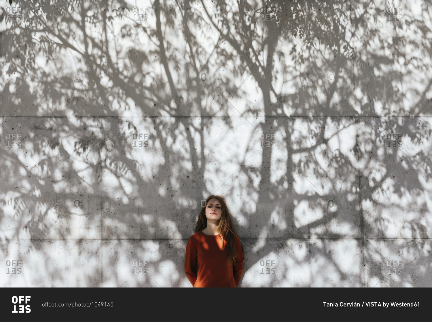 Young woman with hands behind back standing against tree shadow wall