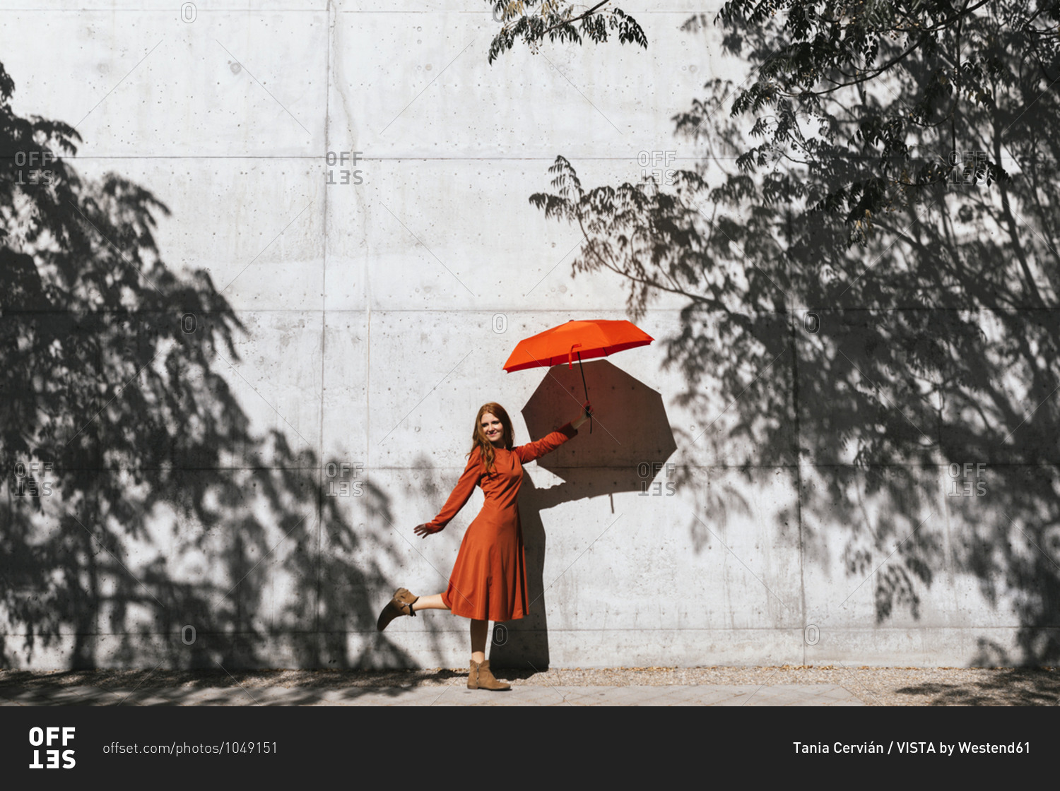 Smiling woman holding red umbrella while standing against tree shadow wall