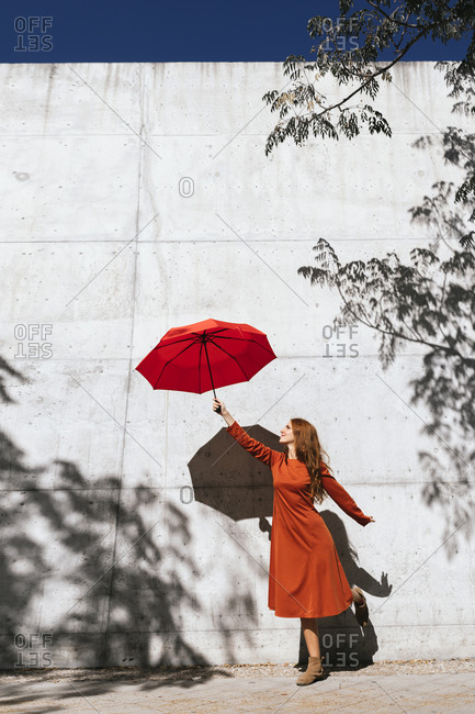 Young woman in red dress holding flying umbrella while standing against tree shadow wall