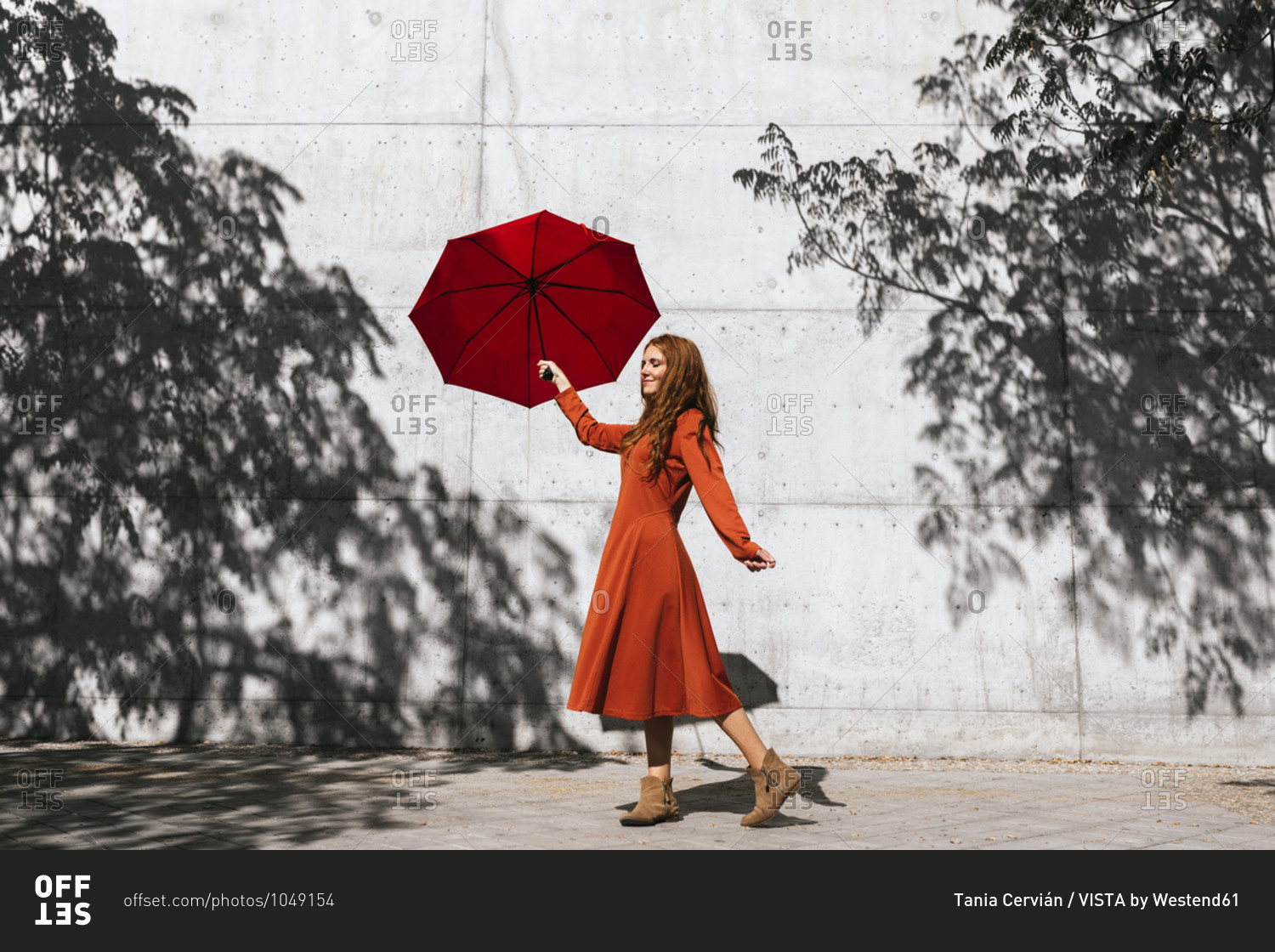 Woman in red dress holding red umbrella while standing against tree shadow wall