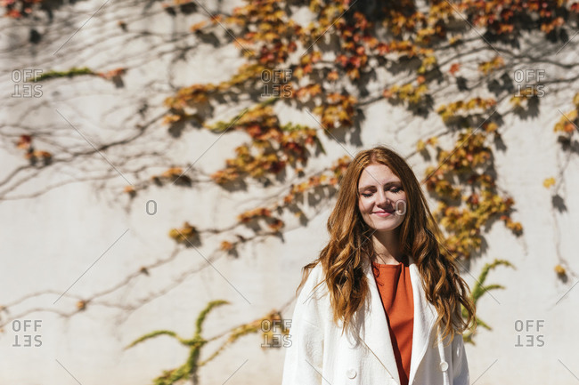 Smiling young redhead woman while standing against ivy wall