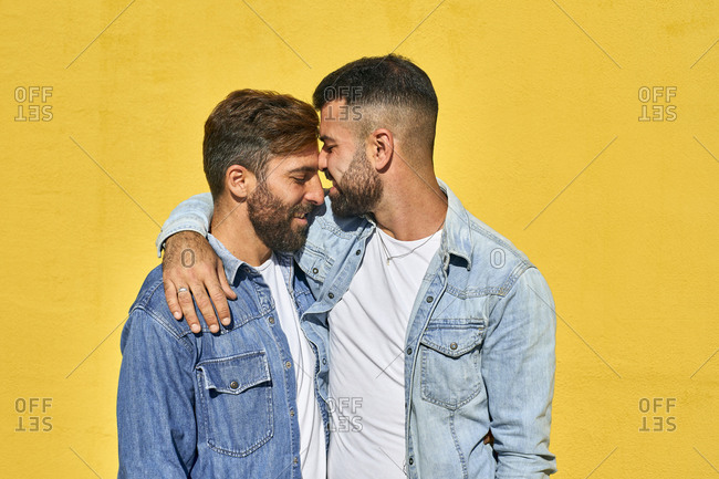 Smiling gay couple standing with arm around standing against yellow wall