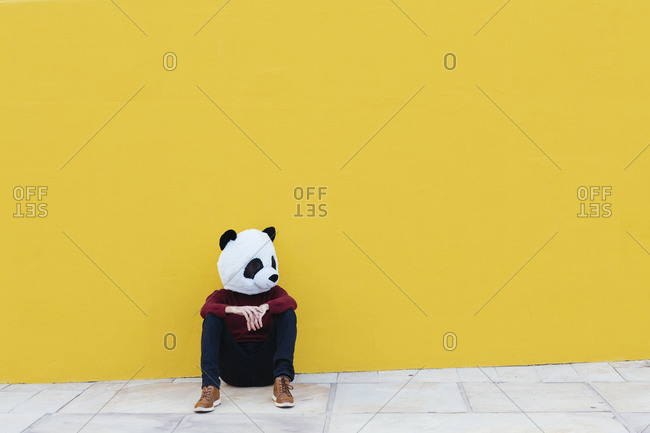 Man wearing panda mask while sitting against yellow wall
