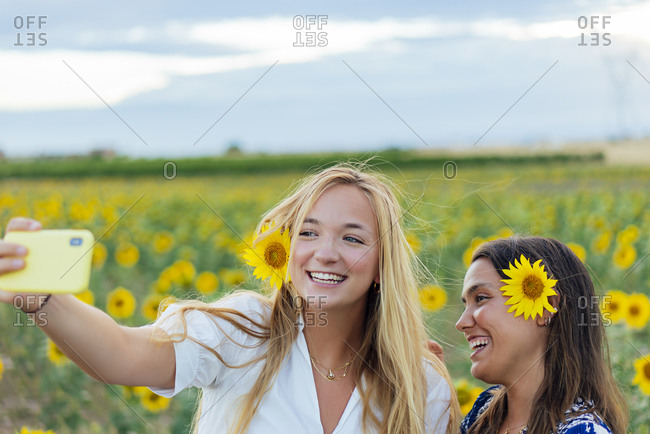 A couple of attractive young women one blonde and the other brunette posing in their designer dresses in a field of sunflowers using their smartphone