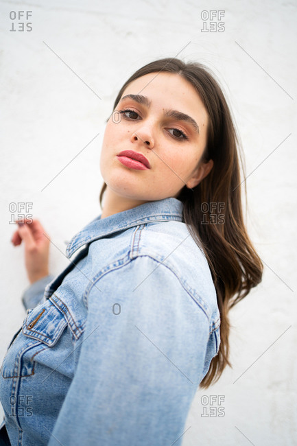 A stylish young woman wearing a light blue jean jacket looking at camera in front of light background