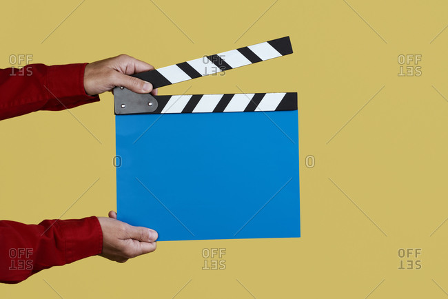 Closeup of a man wearing a red shirt about to clap a blue clapperboard on an orange background
