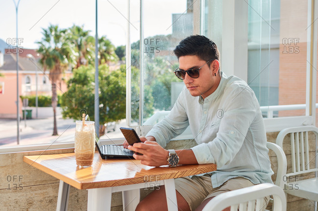 Side view of young stylish male freelancer using smartphone while working remotely on laptop in cafe
