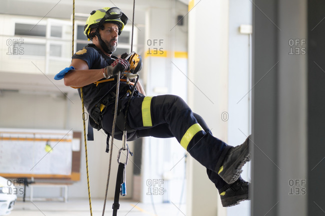 Young fireman in protective hardhat and gloves ascending wall on colorful rope during routine practices