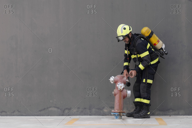 Firefighter in striped uniform and hardhat with extinguisher touching fire hydrant while standing near cement wall on pavement during routine practices