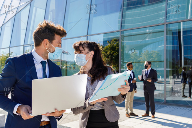 Unrecognizable young man and woman in formal suits and medical masks examining documents while working together on street using laptop near modern business building