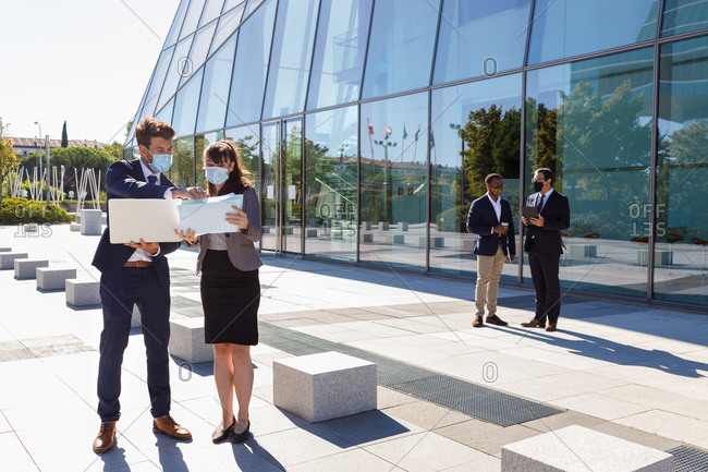 Unrecognizable young man and woman in formal suits and medical masks examining documents while working together on street using laptop near modern business building