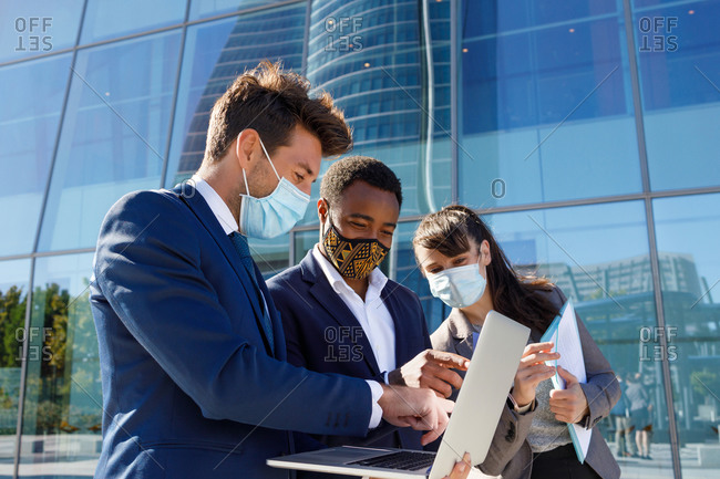 Crop of anonymous diverse coworkers working on laptop in formal suit and medical mask using laptop while standing on street