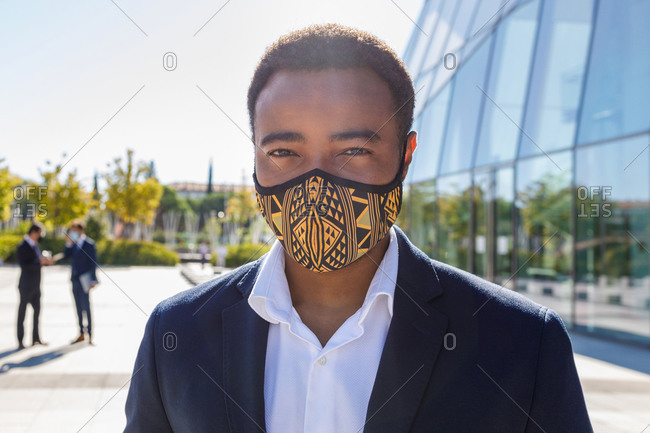 Happy young African American male entrepreneur with mask in classy suit standing on street neat contemporary business building and looking at camera