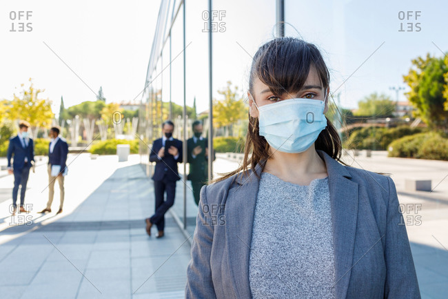 Cheerful young female entrepreneur in elegant outfit looking at camera and medical mask while standing on street near office glass building and anonymous colleagues