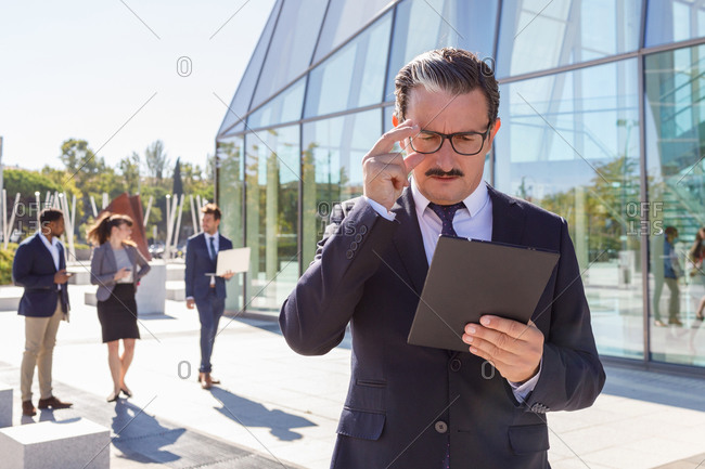 Serious middle aged male manager with mustache in classy outfit holding documents in hand and looking away while thinking about business strategy on street
