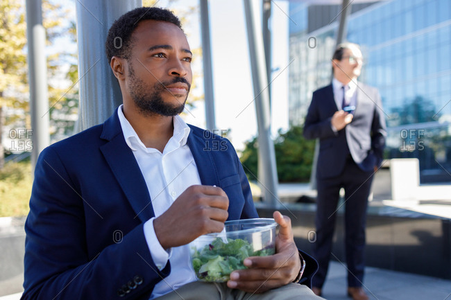 Pensive young black male entrepreneur in formal suit eating healthy takeaway salad while having break on street on sunny day