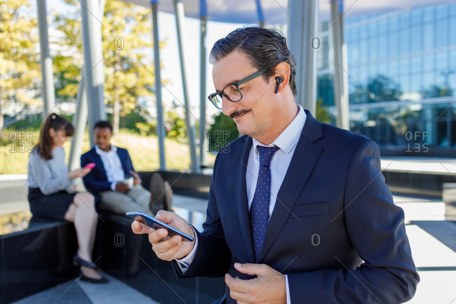 Positive respectable adult businessman in elegant suit and eyeglasses smiling while browsing smartphone on street near modern skyscrapers