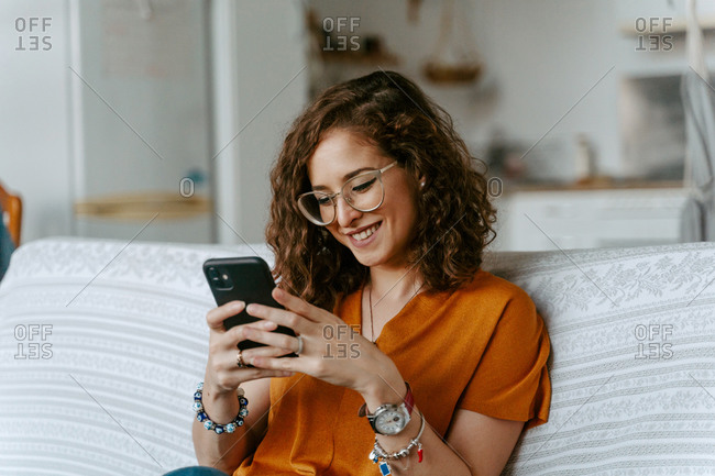 Concentrated young female with curly hair in casual clothing surfing internet while resting on sofa in bright room