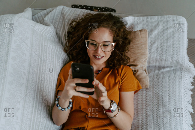 Top view of concentrated young female with curly hair in casual clothing surfing internet while resting on sofa in bright room