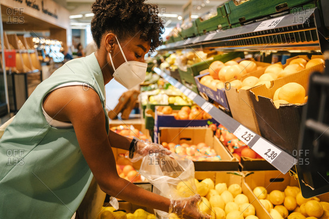 Side view of young African American female in protective mask and gloves choosing red apples while visiting grocery store during coronavirus pandemic