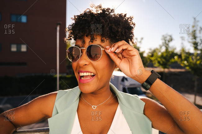 Joyful young African American female with Afro hairdo and trendy sunglasses leaning elbow on railing and looking away while enjoying sunny summer day in park