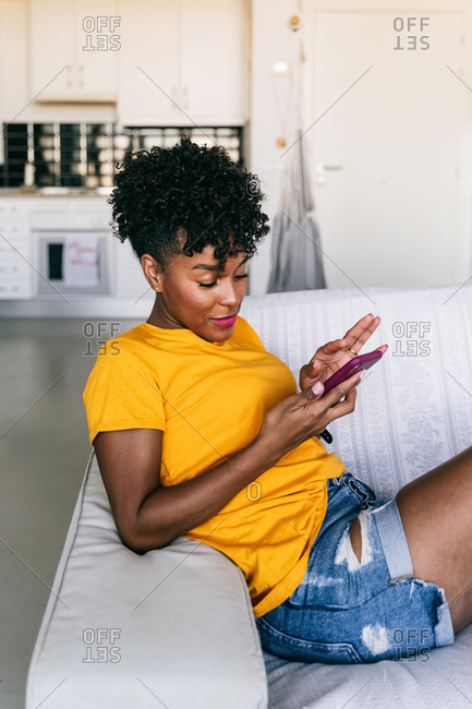 Side view of excited African American millennial female in casual outfit browsing mobile phone and laughing while resting on sofa in modern apartment