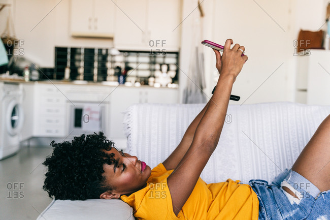 Side view of excited African American millennial female in casual outfit taking selfie with mobile phone and laughing while resting on sofa in modern apartment