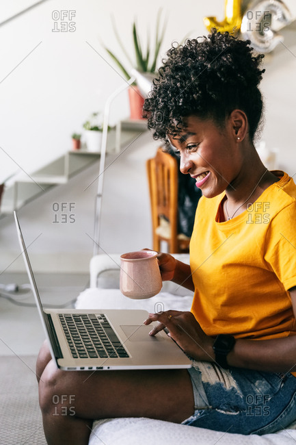 Focused young African American female freelancer with cup of coffee in hand sitting on sofa and browsing laptop while working remotely on project at home