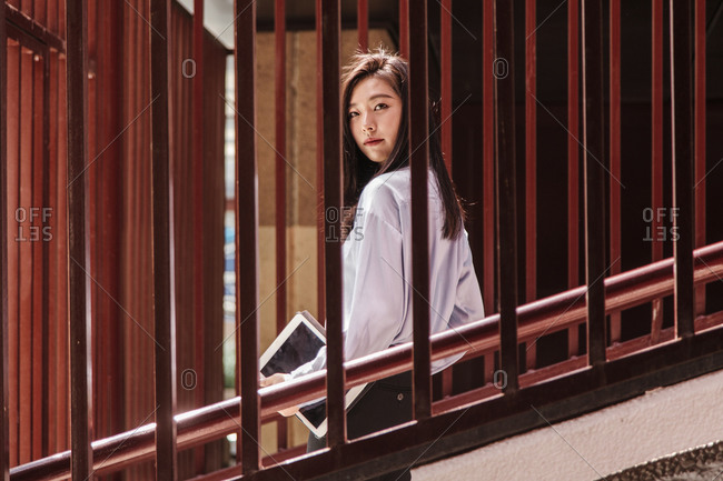 Side view of young attentive woman with long dark hair in classy outfit walking downstairs with hands in pocket and looking at camera through metal railings