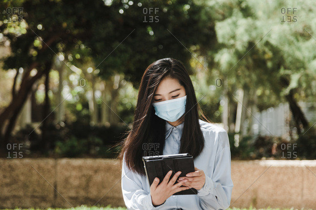 Attentive young Asian female freelancer in formal clothes and medical mask working on tablet while sitting in green park on sunny day