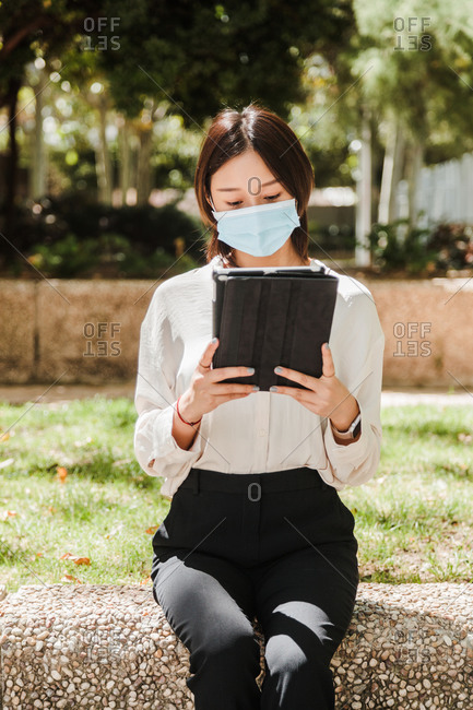 Attentive young Asian female freelancer in formal clothes and medical mask working on tablet while sitting in green park on sunny day