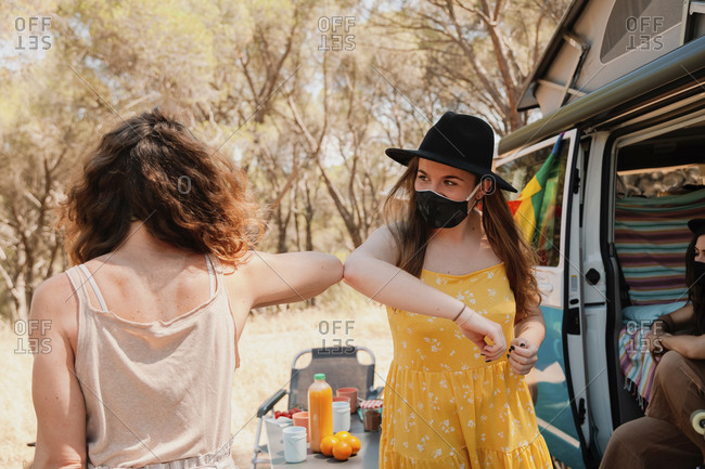 Friendly women in protective masks standing near van in forest and bumping elbows while greeting each other during coronavirus epidemic