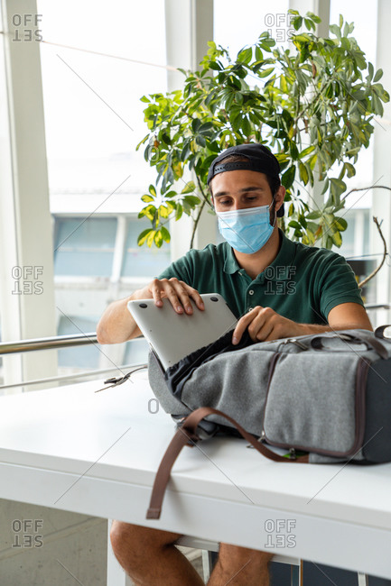 Traveling male in medical mask sitting at table in departure lounge of airport and putting laptop in backpack while waiting for flight during coronavirus epidemic
