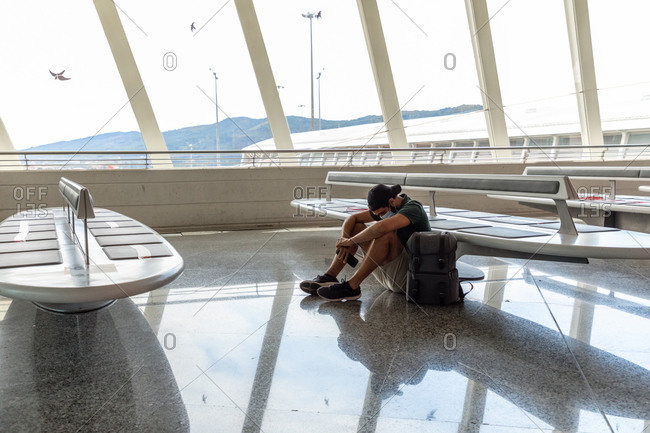 Side view of discouraged male tourist in protective mask sitting on floor in departure lounge of airport with phone while waiting for flight during coronavirus pandemic