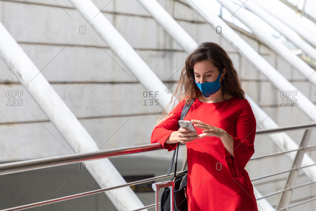 Female tourist in protective mask standing with luggage along hall in airport and reading messages on smartphone while waiting for flight during coronavirus pandemic