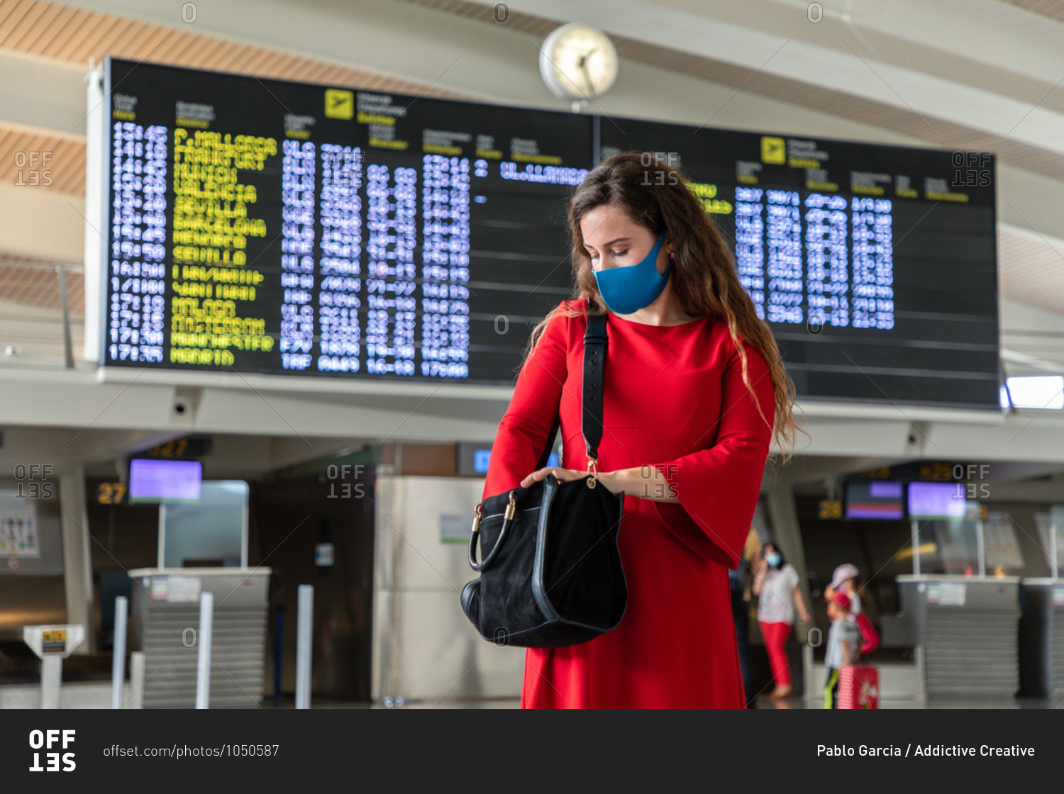 Female tourist wearing protective mask standing in airport against departure board and checking bag while waiting for flight during coronavirus epidemic