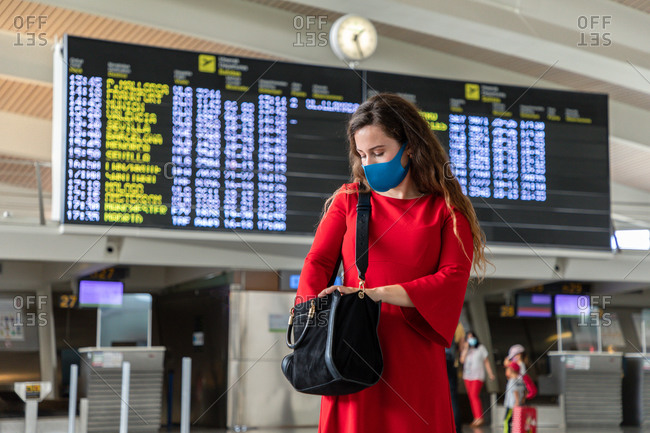 Female tourist wearing protective mask standing in airport against departure board and checking bag while waiting for flight during coronavirus epidemic