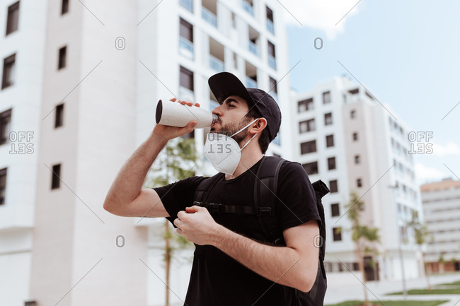 Low angle of male in respiratory mask and black wear drinking water from bottle near buildings and looking away