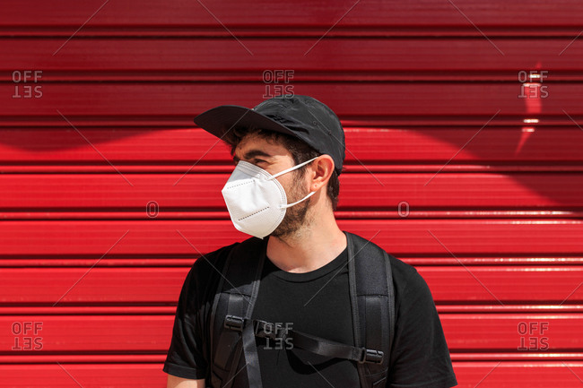 Unrecognizable trendy guy in black apparel on respiratory mask while looking away near colorful wall during coronavirus pandemic