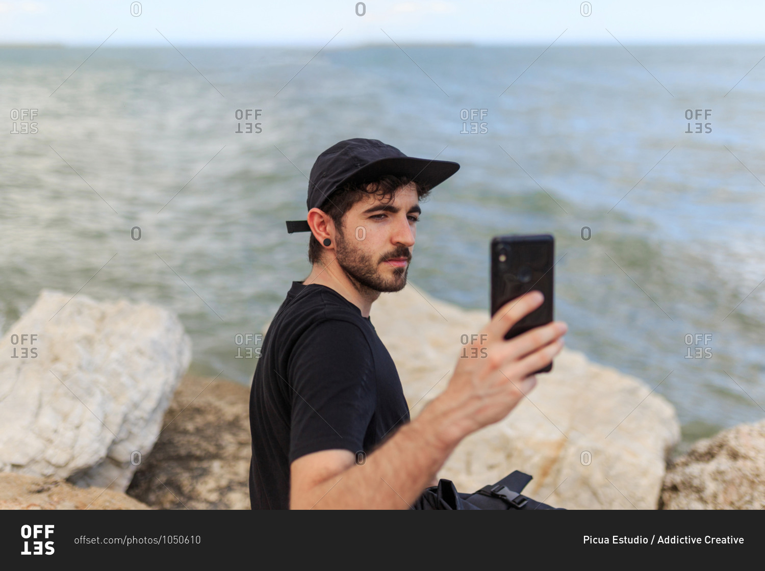 From above side view of bearded male in cap and black clothes taking photo with cellphone while sitting on rough stones near ocean