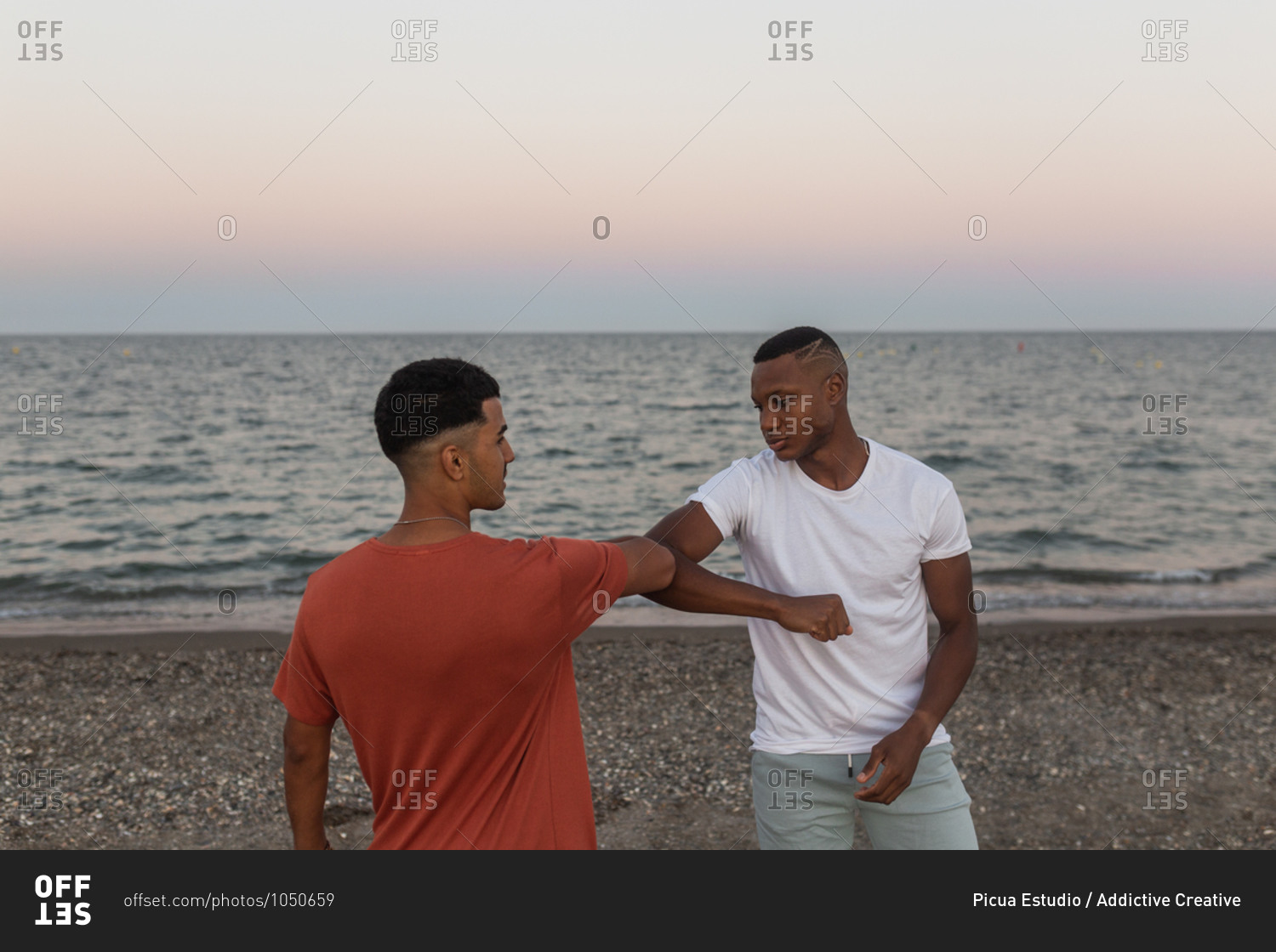 Young trendy African American male partners with modern haircuts greeting each other while bumping elbows on beach near wavy ocean at sunset