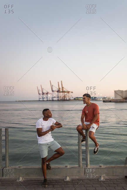 Full body of smiling young ethnic friends spending time together on river embankment in evening