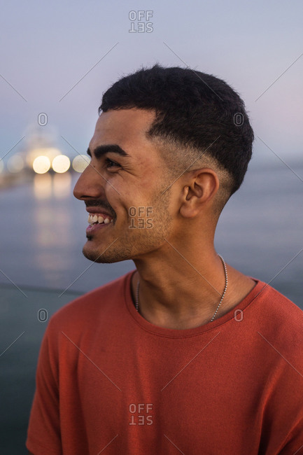 Side view close up portrait of young happy ethnic male traveler standing behind rippled sea under sky in twilight