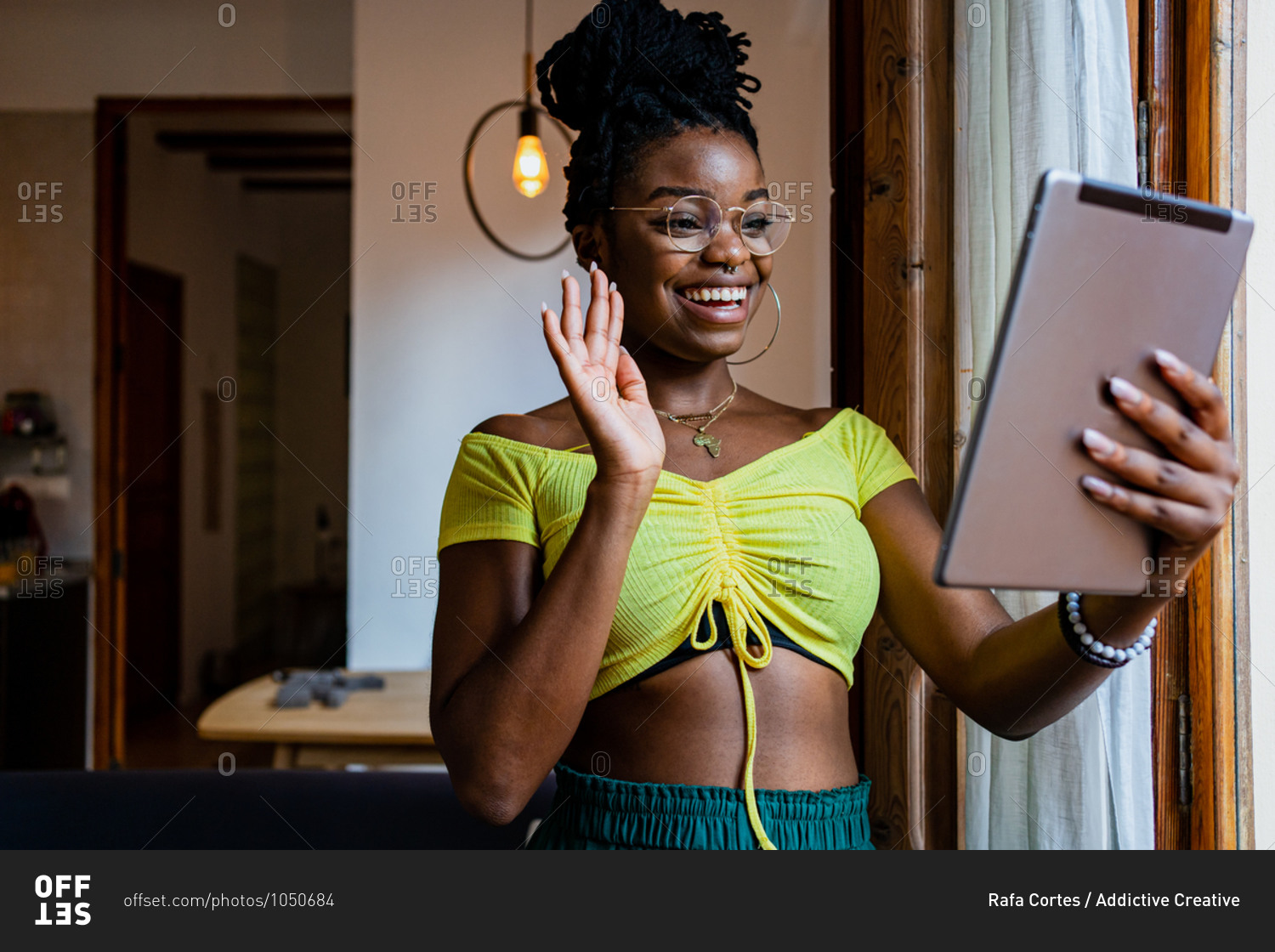 Cheerful young black female in crop top and eyeglasses waving hand and smiling while having video chat on tablet at home