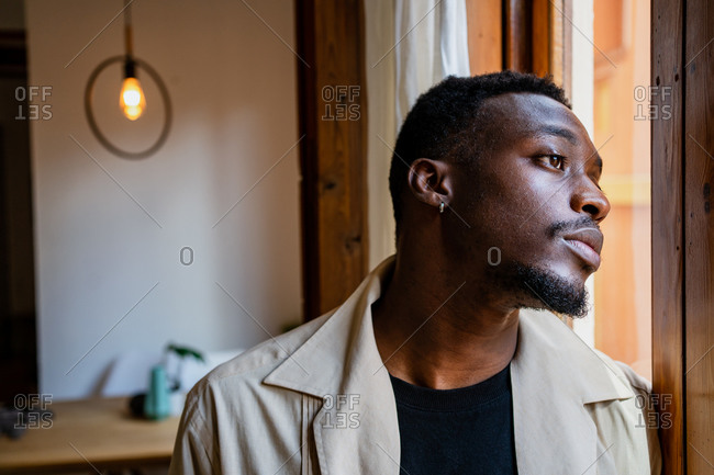 Pensive young African American guy in casual clothes looking out window while resting at home in daylight