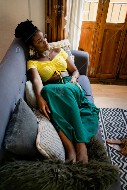 Positive young African American female in bright yellow shirt and transparent eyeglasses looking at camera with bright friendly toothy smile while relaxing on couch in bright studio apartment