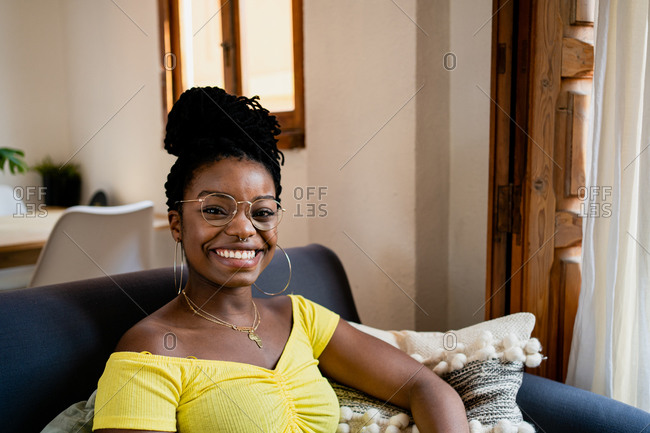 Positive young African American female in bright yellow shirt and transparent eyeglasses looking at camera with bright friendly toothy smile while relaxing on couch in bright studio apartment