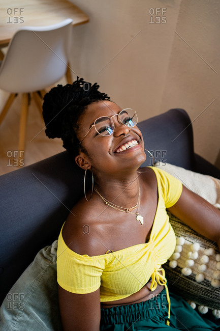 Positive young African American female in bright yellow shirt and transparent eyeglasses looking away with bright friendly toothy smile while relaxing on couch in bright studio apartment